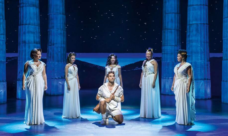 Disneys Hercules singend auf dem Knie im Vordergrund, Saenger in weissen Kleidern im Kreis herumstehend | © Stage Entertainment/Johan Persson 