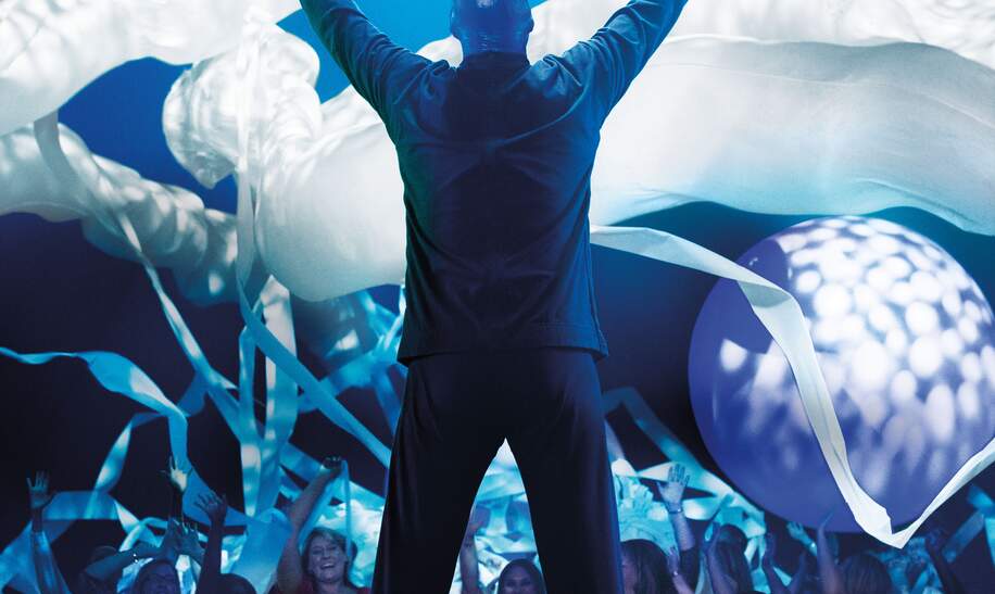 Einer der Kuenstler guckt zum Publikum und hebt die Haende, ueber dem Publikkum schweben Ballons | © Stage Entertainment