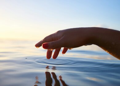  Hand berührt die Wasseroberfläche bei Sonnenuntergang oder -aufgang. | © Unsplash/Yoann Boyer
