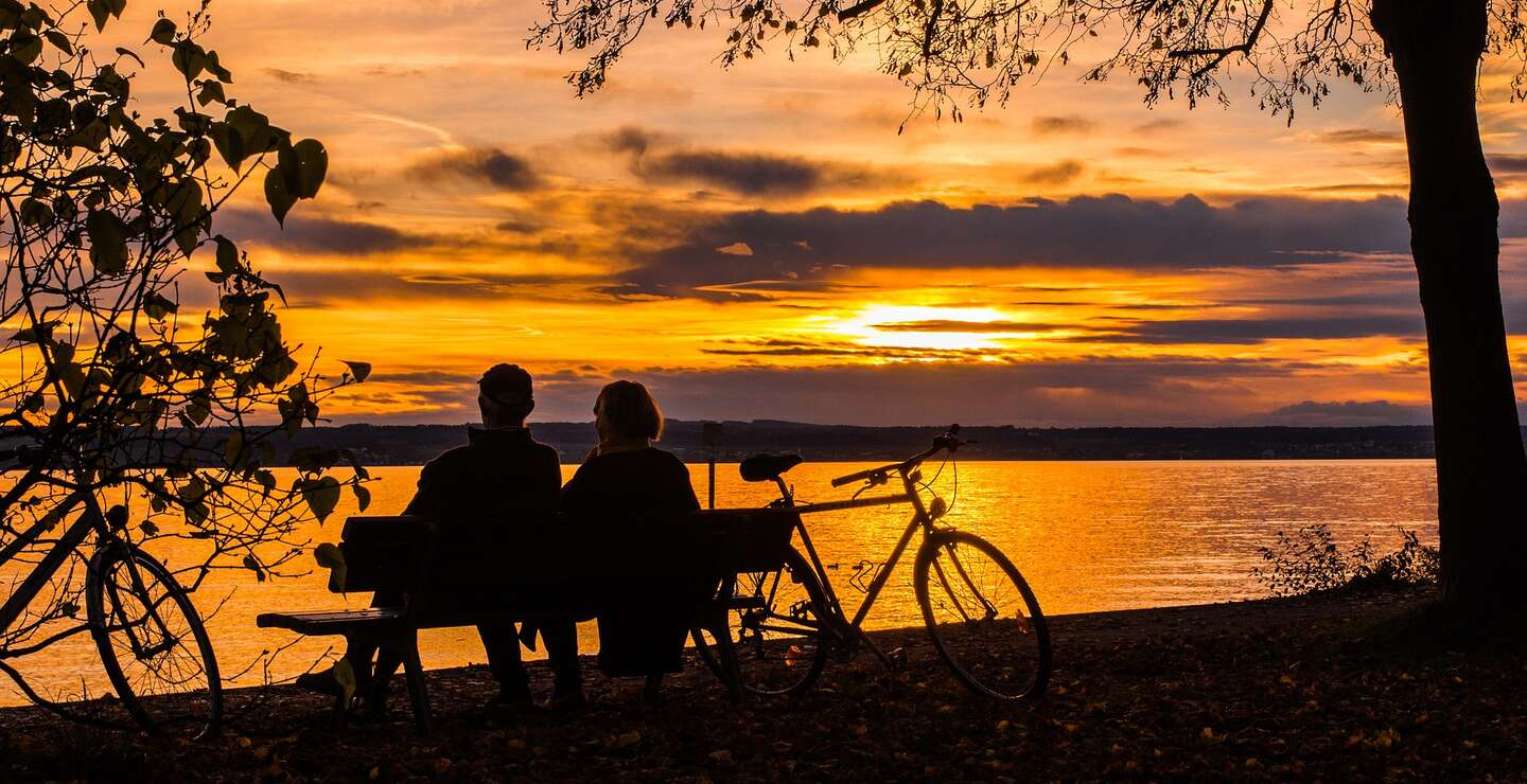 Ein Parr macht eine Pause vom radgahren und setzt sich auf eine Bank in der Naehe des Sees,um einen schoenen Sonnenuntergang zu beobachten | © Pixabay.com/blickpixel 