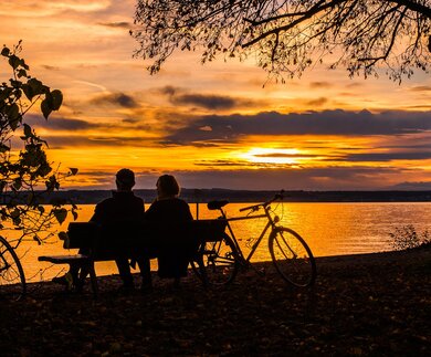 Ein Parr macht eine Pause vom radgahren und setzt sich auf eine Bank in der Naehe des Sees,um einen schoenen Sonnenuntergang zu beobachten | © Pixabay.com/blickpixel 