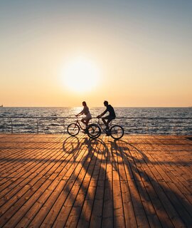 Ein junges Paar faehrt zusammen Fahrrad auf einem Holddeck am Strand bei Sonnenaufgang  | © GettyImages.com/IB_photo