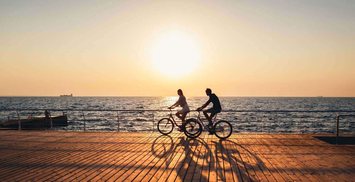 Ein junges Paar faehrt zusammen Fahrrad auf einem Holddeck am Strand bei Sonnenaufgang  | © GettyImages.com/IB_photo