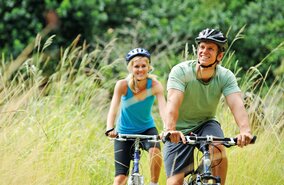 Paar faehrt mit kurzer Kleidung im Feld mit Hohen Graesern und Wald im Hintergrund Fahrrad | © GettyImages.com/warrengoldswain