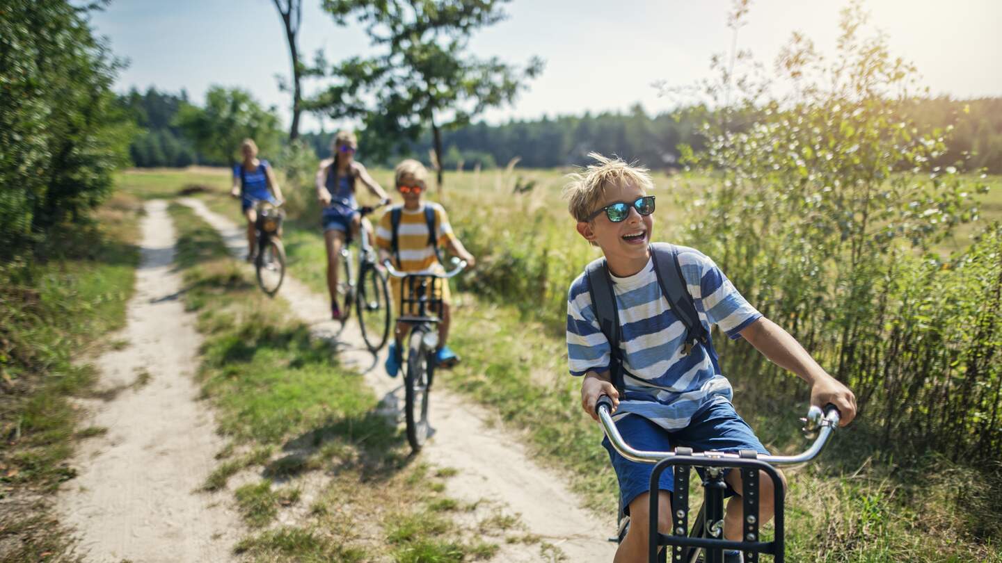 Drei Kinder fahren voraus auf dem Feldweg angrenzed an einen Wald und die Mutter ist verschwommen im Hintergrund zu sehen | © GettyImages.com/Imgorthand