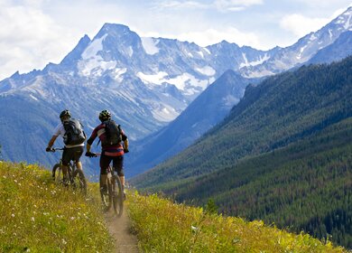Zwei Radfahrer fahren ueber einen Berg in British Columbia, Kanada und sind dabei von hinten zu sehen, vor ihnen liegt ein verschneiter Berg | © GettyImages.com/GibsonPictures