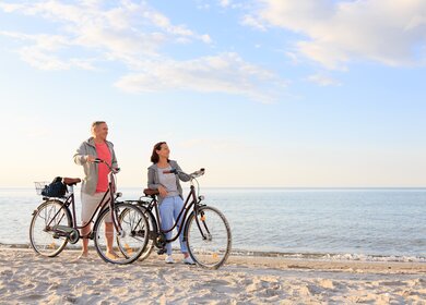 Ein Paar steht mit Fahrraedern am Sandstrand einer Kueste mit blauem Wasser und hellem Himmel | © GettyImages.com/FredFroese