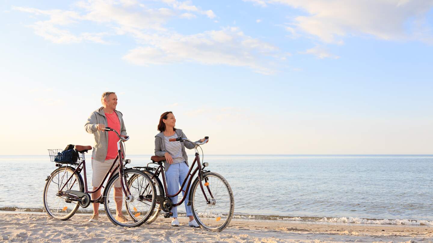 Ein Paar steht mit Fahrraedern am Sandstrand einer Kueste mit blauem Wasser und hellem Himmel | © GettyImages.com/FredFroese