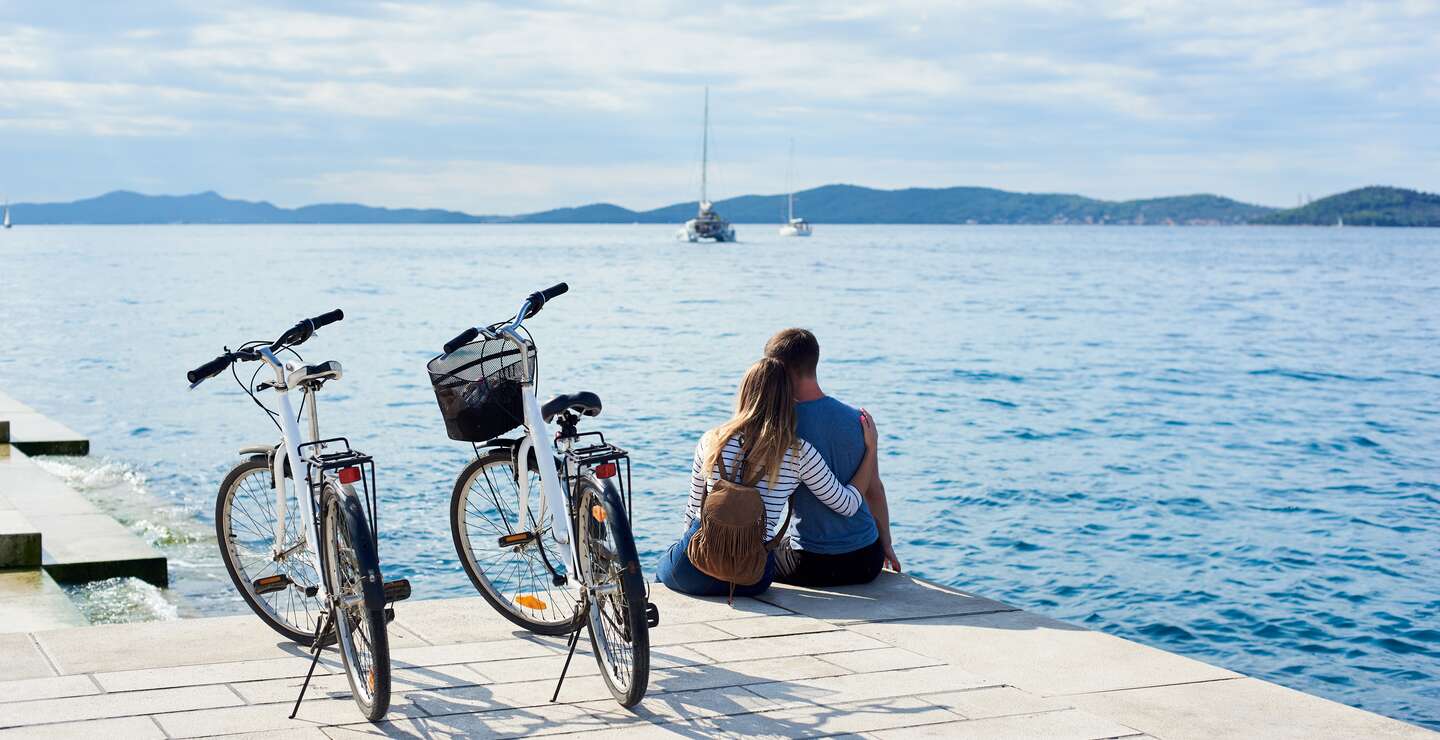 Man und Frau mit Rucksack sitzen umarmt auf Fahrraedern, beobachten schwimmende Kreuzfahrtschiffe in klaren blauen Meer.  | © Gettyimages.com/anatoliy_gleb