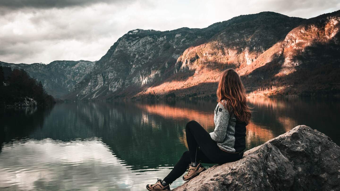 Eine Frau die auf einem grossen Felsen am Rand sitzt und auf eine Bergenlandschaft blickt | © Pixabay.com/sallycadore