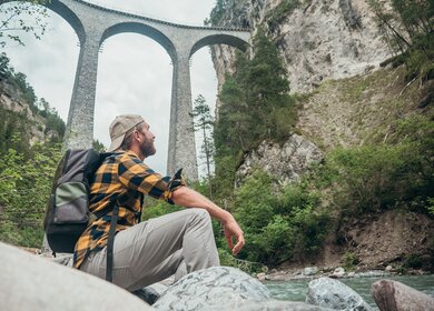 Ein Mann sitzt auf Felsen am Fluss und schaut auf eine grosse Steinbogenbruecke in der Ferne | © Gettyimages.com/Mystockimages