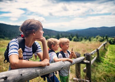 Drei Kinder im Alter von 10 und 7 Jahren vergnügen sich beim Wandern auf den Feldern und Wiesen. Sanft geneigte Huegel, die von Mischwald bedeckt sind, sind im Hintergrund zu sehen. Kinder lehnen sich an einen Holzzaun.  | © Gettyimages.com/Imgorthand