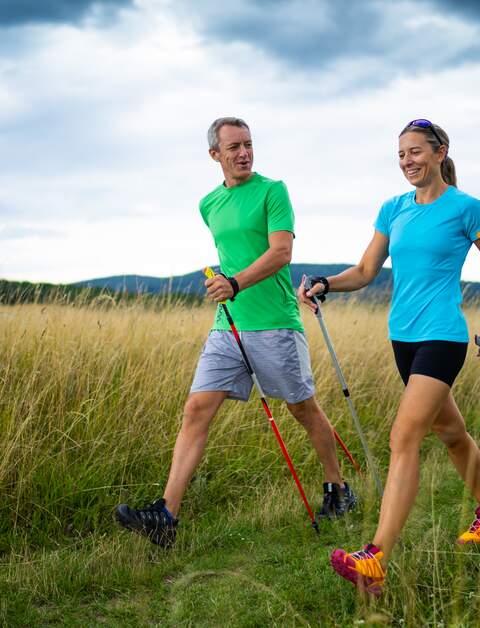 Aktives laechelndes Paar mittleren Alters, das mit skandinavischen Fahrraedern auf einer Wiese mit dunklen Wolken im Vordergrund spazieren geht | © Gettyimages.com/amriphoto