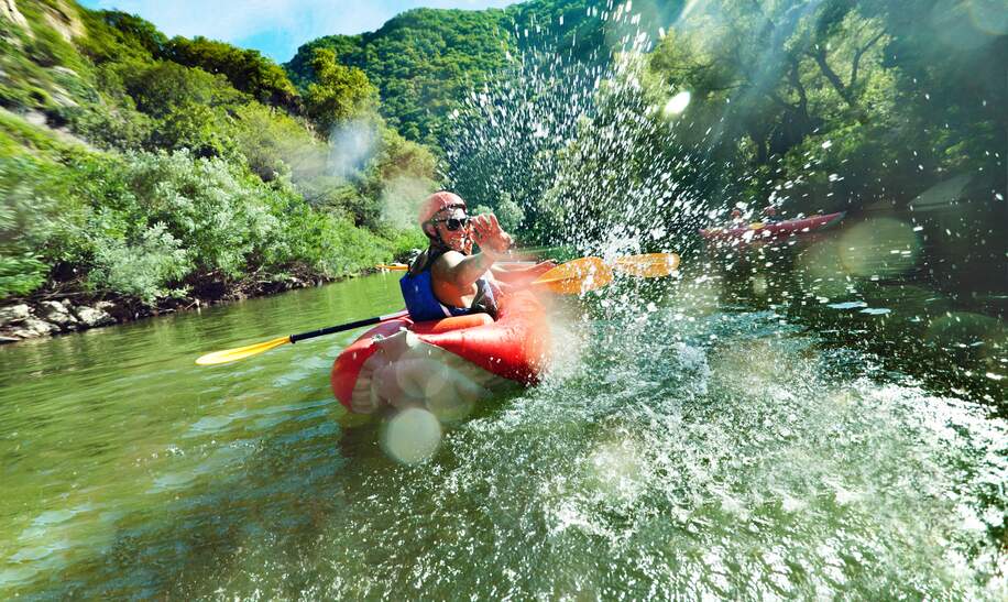 Ein Mann schaut in die Kamera und schwimmt in einem roten aufblasbaren Kanu auf dem ruhigen Wasser eines Flusses | © Gettyimages.com/VILevi