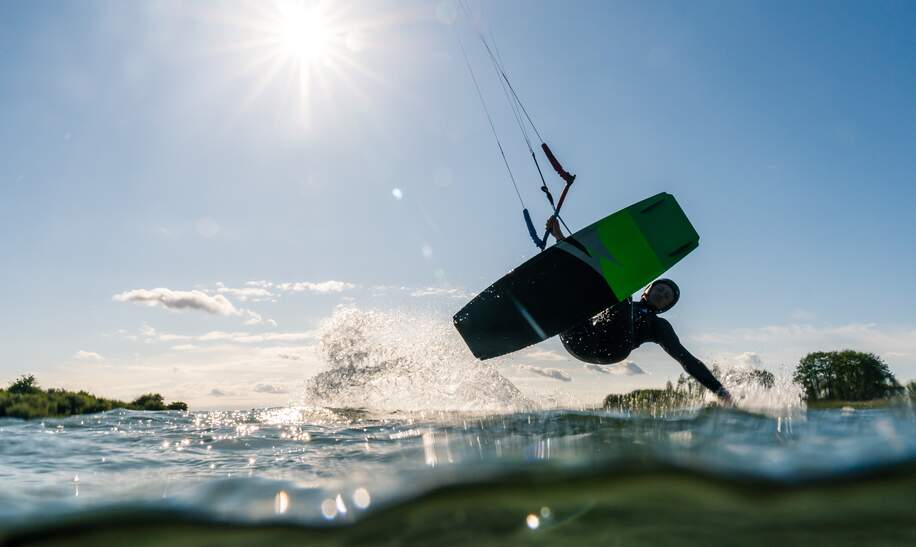 Kitesurfer gleitet mit einer Hand ueber das Wasser, fotografiert von der Wasseroberflaeche aus bei Sonnenschein | © Gettyimages.com/Jens Breuer