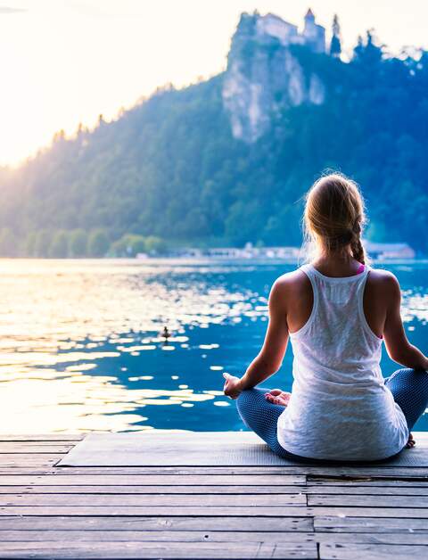 Junge Frau meditiert am Morgen auf einem Holzsteg am Ufer eines Sees, während die Sonne über den bewaldeten Bergen im Hintergrund aufgeht | © Gettyimages.com/microgen