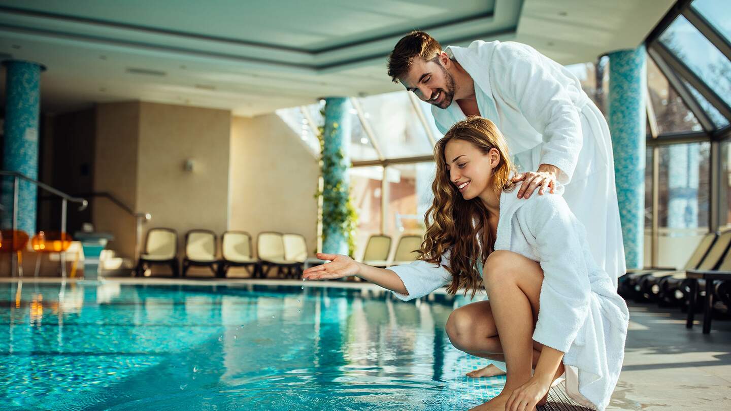 Junge attraktive Frau im Bademantel testet das Wasser des Wellnesspools mit ihrem Freund | © Gettyimages.com/bernardbodo