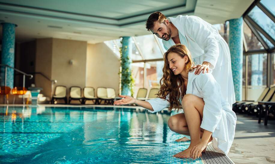Junge attraktive Frau im Bademantel testet das Wasser des Wellnesspools mit ihrem Freund | © Gettyimages.com/bernardbodo