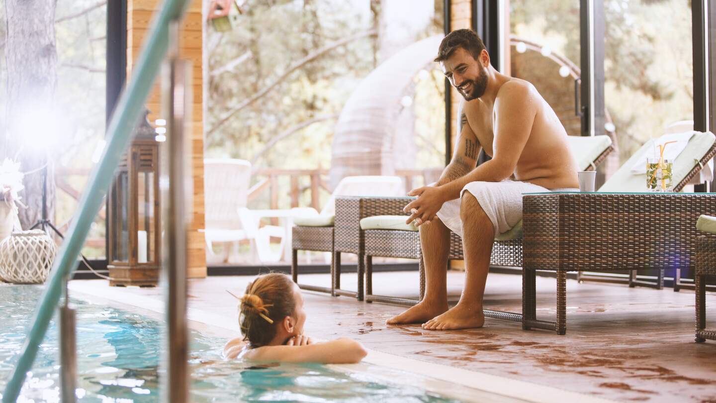 Mann und Frau unterhalten sich im Hallenbad. Die Frau ist in einem Pool und der Mann sitzt auf einem Liegestuhl. | © Gettyimages.com/fotostorm