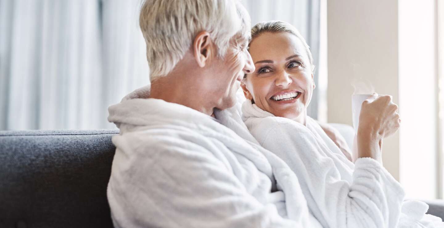 Aufnahme eines froehlichen Paares mittleren Alters, das sich entspannt, waehrend es Bademaentel traegt und tagsüber auf einer Couch entsspannt | © Gettyimages.com/PeopleImages