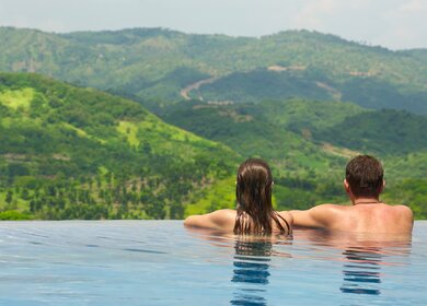 Rueckansicht des Paares mit Blick auf die Berglandschaft vom Infinity-Pool aus | © Gettyimages.com/Soft_Light