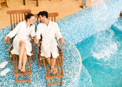 Paar entspannt sich im Bademantel auf Liegestuehlen am Swimmingpool | © Gettyimages.com/fotostorm