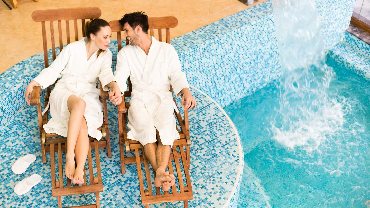 Paar entspannt sich im Bademantel auf Liegestuehlen am Swimmingpool | © Gettyimages.com/fotostorm