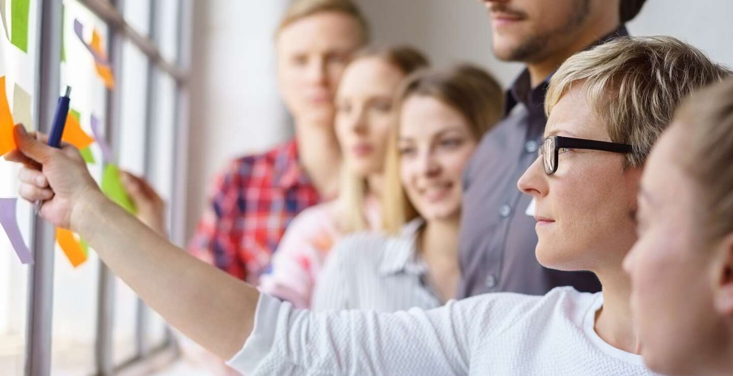 Junges Team von fokussierten Berufseinsteigern, die Ideen diskutieren und mit Haftnotizen an einem Buerofenster brainstormen | © Gettyimages.com/stockfour