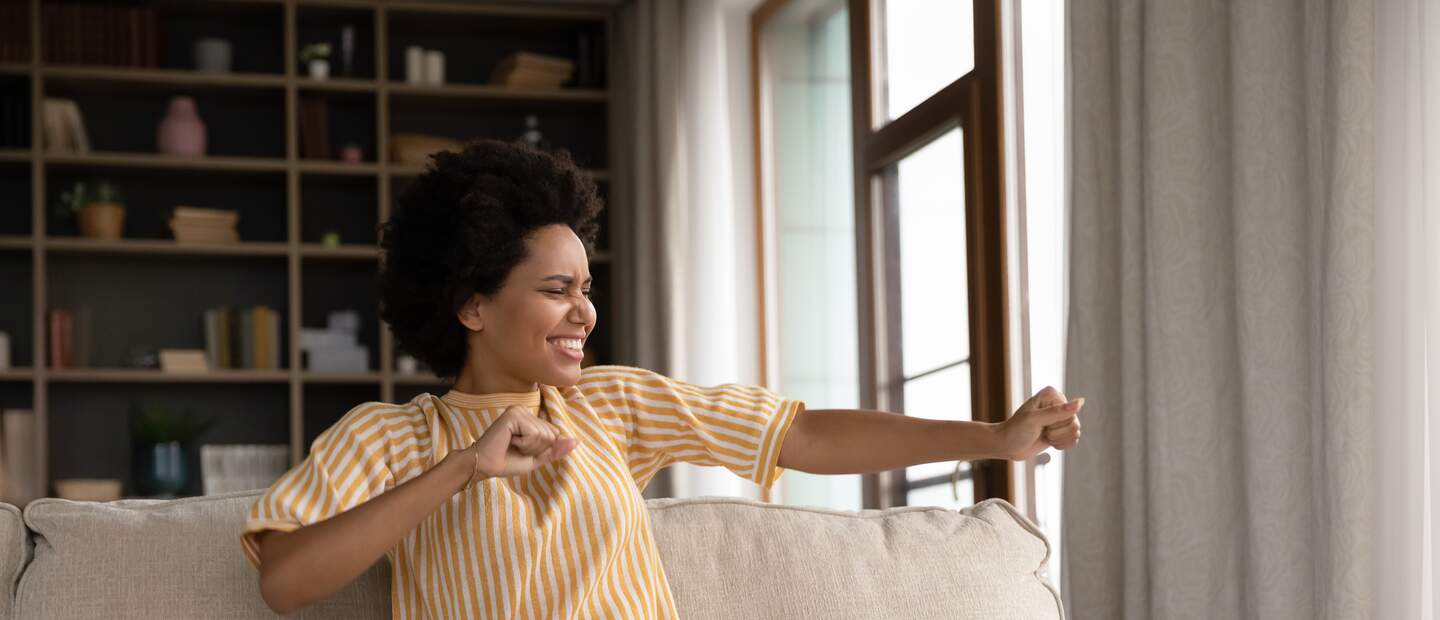  junge afroamerikanische Frau, die zu Hause am Computer arbeitet, den letzten Arbeitstag feiert, Spass allein im Wohnzimmer hat | © Gettyimages.com/fizkes