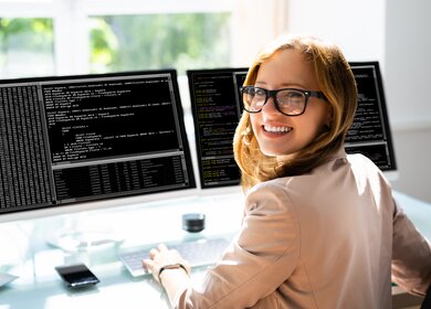 Frau mit Brille schaut laechelnd nach links, während vor ihr zwei Bildschirme stehen auf denen sie programmiert | © Gettyimages/AndreyPopov