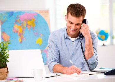 Mann telefoniert mit Kabeltelevon und laechelt waehrend er Notizen in sein Buch eintraegt, neben ihm stehen ein aufgeklappter Laptop und eine Pflanze, hinter ihm sind Weltkate und Weltkugel | © Gettyimages/dima_sidelnikov