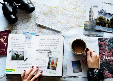 eine flache Anordnung verschiedener Reiseutensilien auf einer Oberflaeche, darunter eine Karte, ein Fernglas, ein Reisepass, Postkarten, Fotos und eine Tasse Kaffee | © gettyimages.com/Rawpixel Ltd.