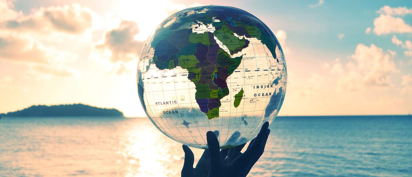 Eine Hand hält eine durchsichtige Weltkugel vor einem Sonnenuntergang am Strand | © Gettyimages.com/jaminwell