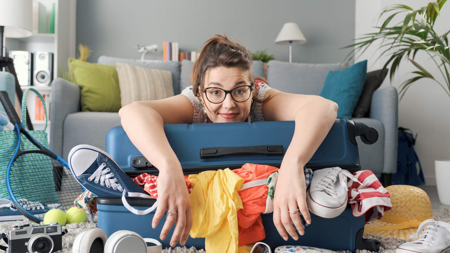Glueckliche junge Frau packt fuer eine Reise, sie lehnt sich an ihren vollen Trolley-Koffer und laechelt | © Gettyimages.com/demaerre