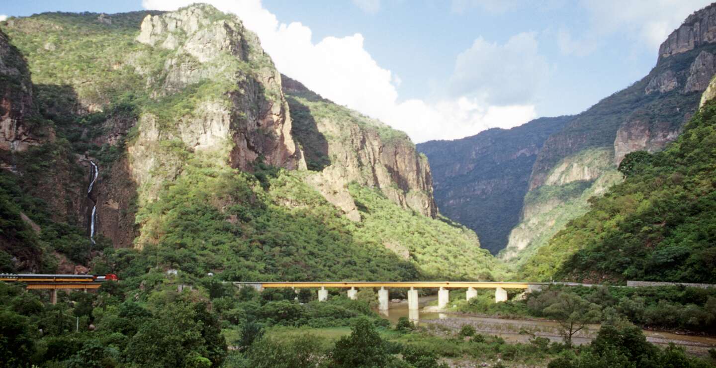 Der  Zug CHEPE fährt über eine Brücke im Kupfercanyon im Norden Mexikos im Hintergrund die bewaldete Bergkulisse | © Native Trails