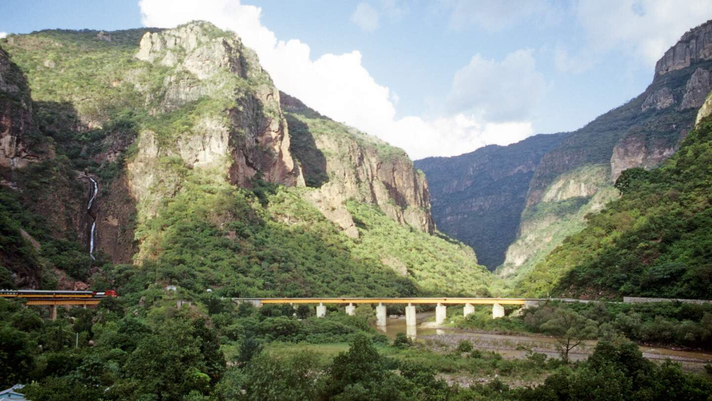 Der  Zug CHEPE fährt über eine Brücke im Kupfercanyon im Norden Mexikos im Hintergrund die bewaldete Bergkulisse | © Native Trails